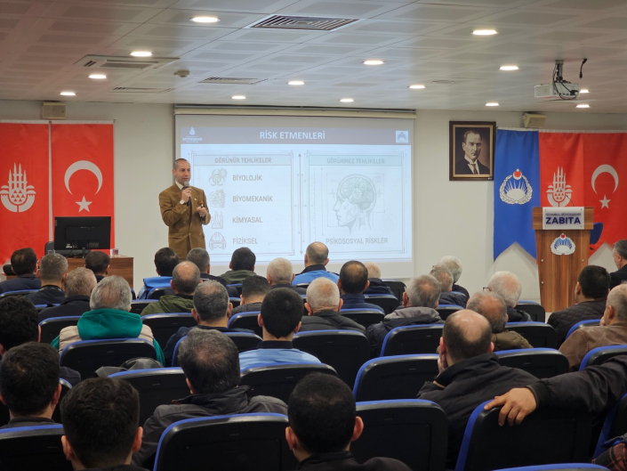Bir konferans salonunda, sahnede "Risk Etmenleri" başlıklı slaytın önünde konuşan kahverengi ceketli eğitmen ve onu dinleyen kalabalık zabıta personeli.