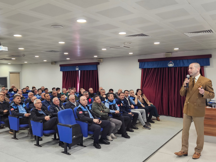 Bir konferans salonunda, kahverengi ceketli bir eğitmen ayakta mikrofona konuşurken mavi koltuklarda oturan üniformalı zabıta personelleri onu dinliyor.