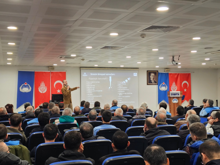 Bir konferans salonunda, sahnede "Stresin bireysel sonuçları: Fizyolojik Sorunlar" başlıklı slayt önünde anlatım yapan eğitmen ve eğitimi dinleyen zabıta personelleri.
