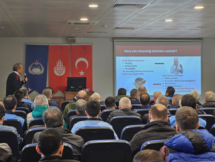 Bir eğitmen, konferans salonundaki katılımcılara "Hava yolu tıkanıklığı belirtileri nelerdir?" başlıklı bir ilkyardım eğitimi veriyor.