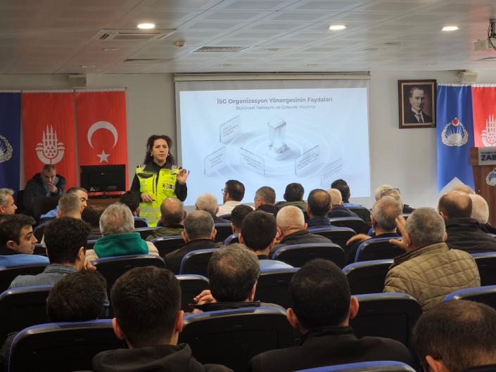 Bir konferans salonunda, fosforlu ikaz yeleği giymiş kadın eğitmen "İSG Organizasyon Yönergesinin Faydaları" başlıklı slayt önünde sunum yaparken kalabalık dinleyici kitlesi onu izliyor.