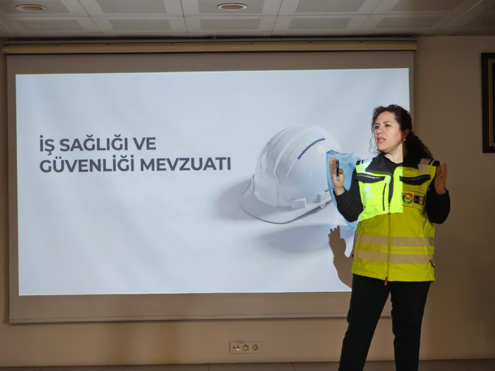 "İŞ SAĞLIĞI VE GÜVENLİĞİ MEVZUATI" başlıklı sunum perdesinin önünde anlatım yapan fosforlu ikaz yelekli kadın eğitmen.
