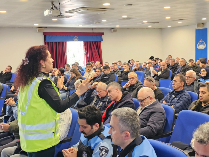 Bir konferans salonunda, fosforlu yeşil ikaz yeleği giymiş bir kadın eğitmen ayakta dinleyicilere hitap ediyor.