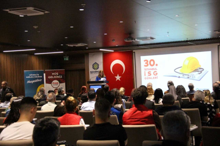 Bir konferans salonunda gerçekleştirilen "30. İstanbul İSG Günleri" etkinliğinin genel görünümü. Kürsüdeki konuşmacı sunum yaparken, arkasındaki projeksiyon perdesinde sarı baret ve stetoskop görseli içeren etkinlik slaytı yer alıyor; ön planda ise dinleyiciler oturmaktadır.
