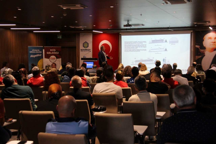 Geniş bir konferans salonunda sunum yapan takım elbiseli bir erkek konuşmacı. Kürsünün yanında, büyük bir Türk bayrağı önünde ayakta durarak arkasındaki projeksiyon perdesinde yer alan teknik mimari çizimleri ve mimari detay paragraflarını dinleyicilere açıklamaktadır.