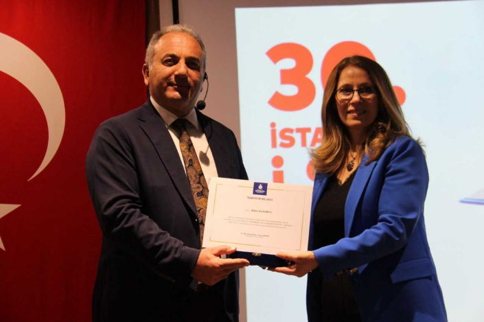 Büyük bir Türk bayrağı ve "30. İstanbul İSG Günleri" yansıtılmış projeksiyon perdesi önünde, saks mavisi ceketli ve gözlüklü bir kadının, takım elbiseli bir erkeğe teşekkür belgesi takdim ettiği anın yakın çekim fotoğrafı.