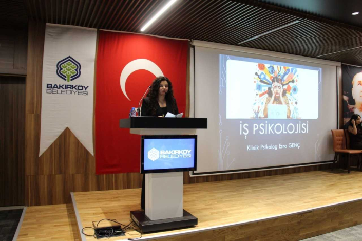 Bakırköy Belediyesi yazan ekranlı kürsüde, büyük bir Türk bayrağı önünde sunum yapan bir kadın konuşmacı. Arkasındaki büyük ekranda stresli bir çalışan illüstrasyonu ile "İş Psikolojisi - Klinik Psikolog Esra Genç" yazılı sunum slaytı yer alıyor.
