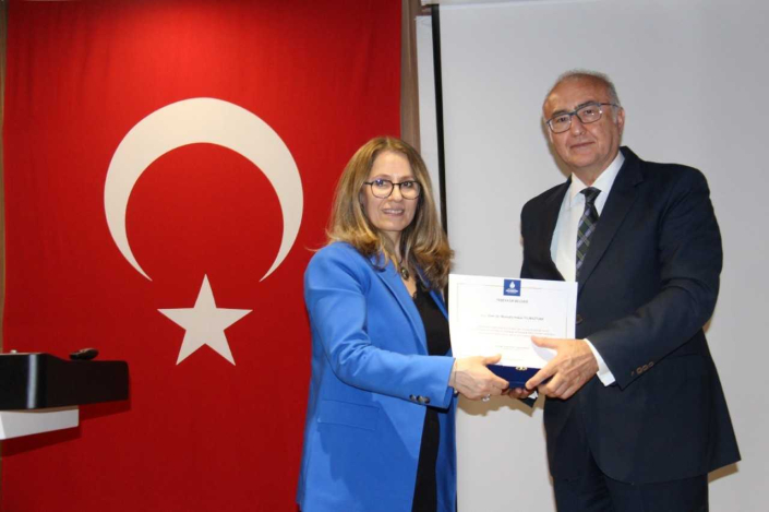 Büyük bir Türk bayrağı önünde, saks mavisi ceketli ve gözlüklü bir kadının, takım elbiseli ve gözlüklü bir erkeğe lacivert kutu üzerinde duran bir teşekkür belgesi takdim ettiği anın yakın çekim fotoğrafı.