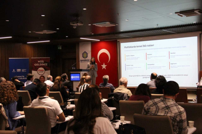 Bir konferans salonunda gerçekleştirilen bir İş Sağlığı ve Güvenliği (İSG) sunumunun genel bir görünümüdür. Sahnedeki konuşmacı, arkasındaki ekranda 'Mutfaklarda temel İSG riskleri' başlığıyla risk faktörlerini listeleyen bir slayt eşliğinde sunum yapmaktadır.