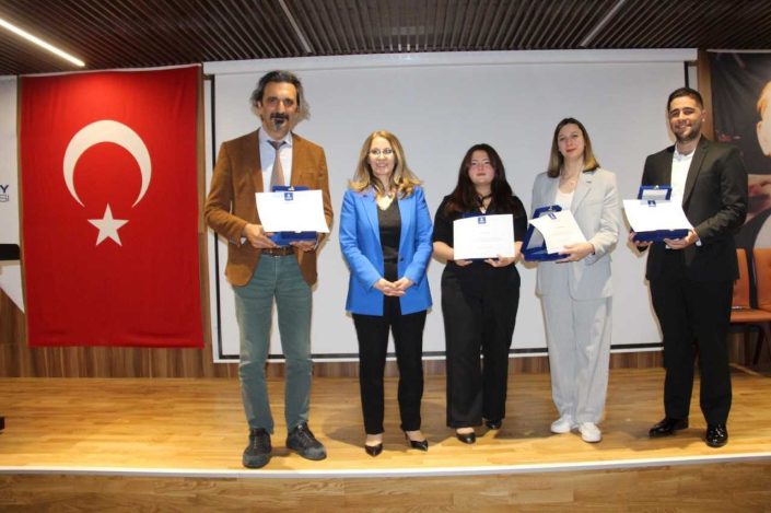 Konferans sahnesinde, büyük bir Türk bayrağı önünde yan yana duran, üçünün elinde mavi plaket kutusu ve başarı belgesi bulunan iki kadın ve iki erkek katılımcı ile ortalarında saks mavisi ceketli bir başka kadın.