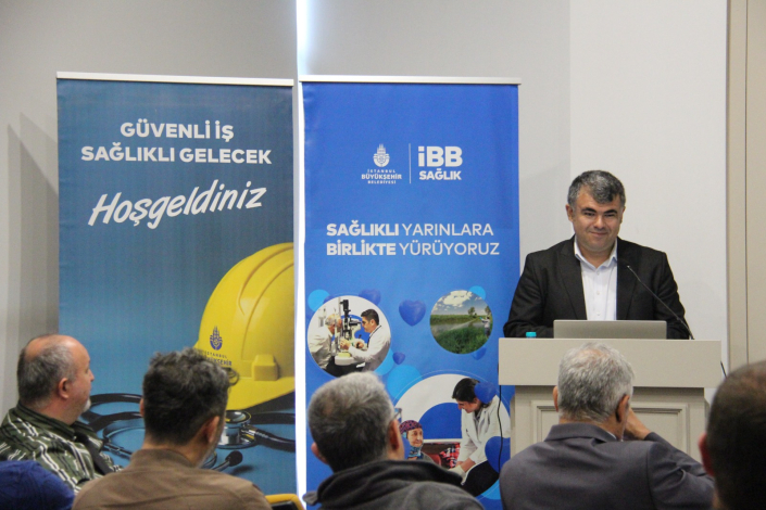 Bir kürsüde konuşma yapan erkek konuşmacı, arkasında "İBB Sağlık" logolu iki adet mavi tanıtım panosu. Panoların üzerinde "Güvenli İş Sağlıklı Gelecek Hoşgeldiniz" ve "Sağlıklı Yarınlara Birlikte Yürüyoruz" yazıları ile sağlık temalı görseller (sarı baret, stetoskop, doktor) yer almaktadır. Ön planda dinleyiciler oturmaktadır.