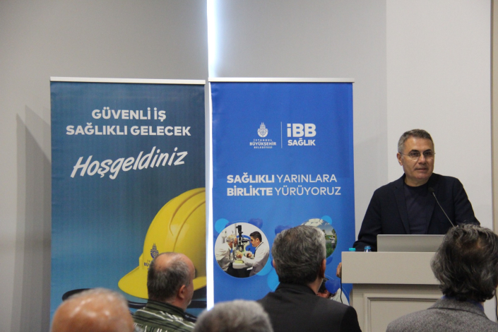 Kürsüde konuşma yapan gözlüklü ve takım elbiseli bir adam ile arka planda "Güvenli İş Sağlıklı Gelecek Hoşgeldiniz" ve İBB Sağlık logolu "Sağlıklı Yarınlara Birlikte Yürüyoruz" yazılı iki mavi tanıtım panosu.
