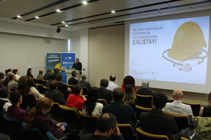 Bir konferans salonunda, üzerinde sarı baret ve stetoskop görseli ile "İBB, Bağlı Kuruluşlar ve İştirakler İSG Koordinasyon Çalıştayı" yazan büyük bir projeksiyon perdesinin önünde konuşma yapan bir sunucu ve onu dinleyen kalabalık bir katılımcı grubu.
