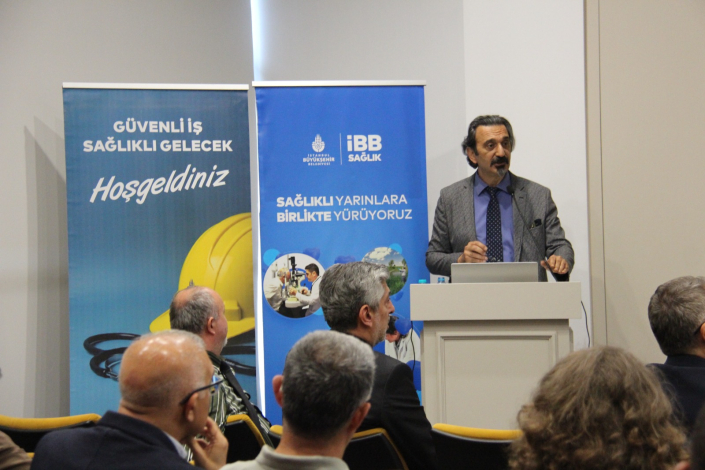 Kürsüde konuşma yapan sunum sahibi ile arkasında "Güvenli İş Sağlıklı Gelecek Hoşgeldiniz" ve "Sağlıklı Yarınlara Birlikte Yürüyoruz" yazılı iki adet İBB Sağlık tanıtım panosu. Ön planda konuşmacıyı dinleyen katılımcıların başları ve arkadan görünümleri yer almaktadır.