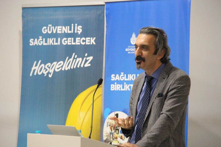 Kürsüde konuşma yapan sunum sahibi, eliyle jest yaparak konuyu anlatmaktadır. Arkasında "Güvenli İş Sağlıklı Gelecek Hoşgeldiniz" yazılı ve İstanbul Büyükşehir Belediyesi amblemli iki adet mavi tanıtım panosu yer almaktadır.