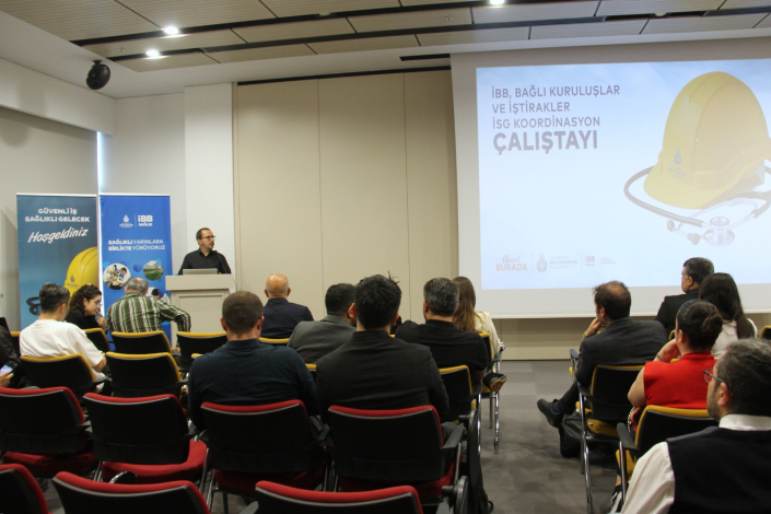 "İBB, Bağlı Kuruluşlar ve İştirakler İSG Koordinasyon Çalıştayı" başlıklı bir sunumun yapıldığı bir konferans salonunun içini gösteren fotoğraf. Sunumda sarı bir baret ve stetoskop resmi yer alıyor. Kürsüde konuşma yapan bir kişi ve onu dinleyen bir dinleyici kitlesi bulunuyor.