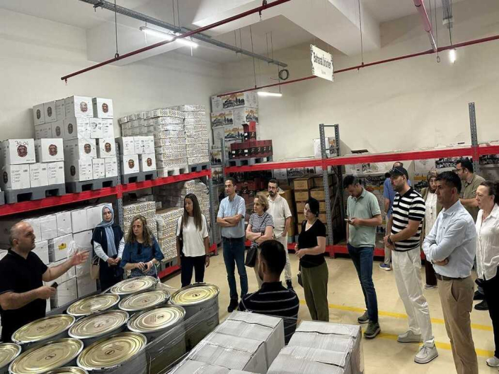 Depolama alanındaki "Baharat Ürünleri" reyonunda, koli ve teneke ambalajların önünde toplanan gruba bir yetkilinin bilgi verdiği saha incelemesi anı.