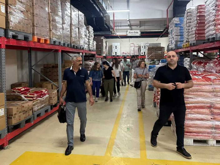 Yüksek raflarla çevrili bir kuru gıda deposunda yerde sarı çizgilerin bulunduğu koridorda yürüyen bir grup insan.