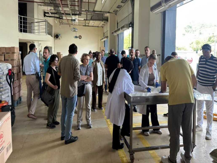 Tesis içerisinde önlüklü personelinde etrafında bulunduğu tezgahta yapılan işlemi izeyen kalabalık bir heyet.