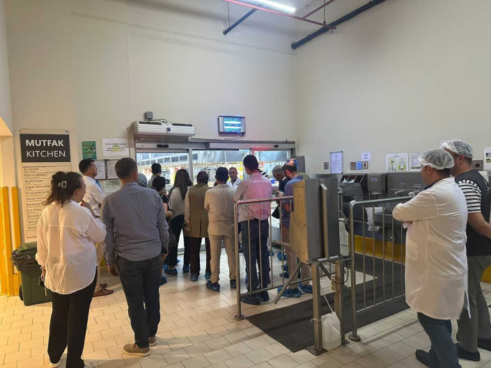 Duvarında "MUTFAK / KITCHEN" tabelası bulunan tesis girişinde, beyaz önlük ve bone giymiş görevliler ve mutfak alanını izleyen katılımcı grubu.