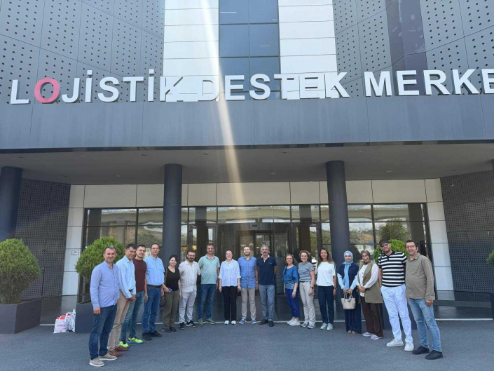Üzerinde büyük harflerle "LOJİSTİK DESTEK MERKEZİ" yazan modern bir binanın girişinde, açık havada yan yana dizilerek poz veren çalışan ve teknik personel.