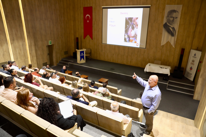 Seminer salonunda, projeksiyon perdesindeki 'Davranış' başlıklı sunum eşliğinde konuyu anlatan eğitmen ve onu izleyen katılımcılar.