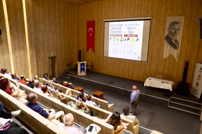 Seminer salonundaki büyük projeksiyon perdesinde bir kitabın İngilizce ve Türkçe kapak görsellerinin yer aldığı sunumu gerçekleştiren eğitmen ve onu izleyen dinleyiciler.