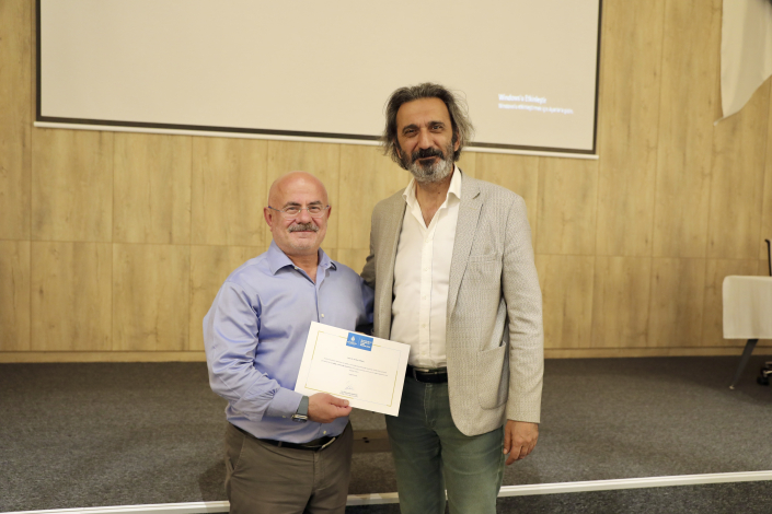 Bir seminer sahnesinin önünde birinin elinde teşekkür belgesi olan ve yan yana durarak poz veren iki erkek katılımcı.