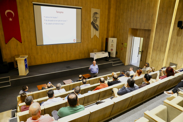 Bir seminer salonunda sahnedeki eğitmenin sunum yaptığı, dinleyicilerin ise sıralarda oturarak eğitimi takip ettiği kurumsal bir eğitim salonu.