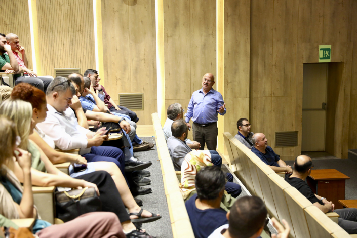 Seminer salonundaki basamaklı sıraların arasında durarak dinleyicilerle soru-cevap veya interaktif konuşma yapan bir eğitmen.