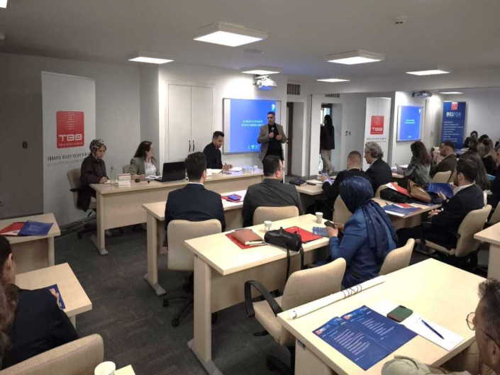 TBB yazan flamalar bulunan salonda yapılan sunumu gerçekleştiren konuşmacı ile masalara torumuş şekilde onu dinleyen katılımcılar.