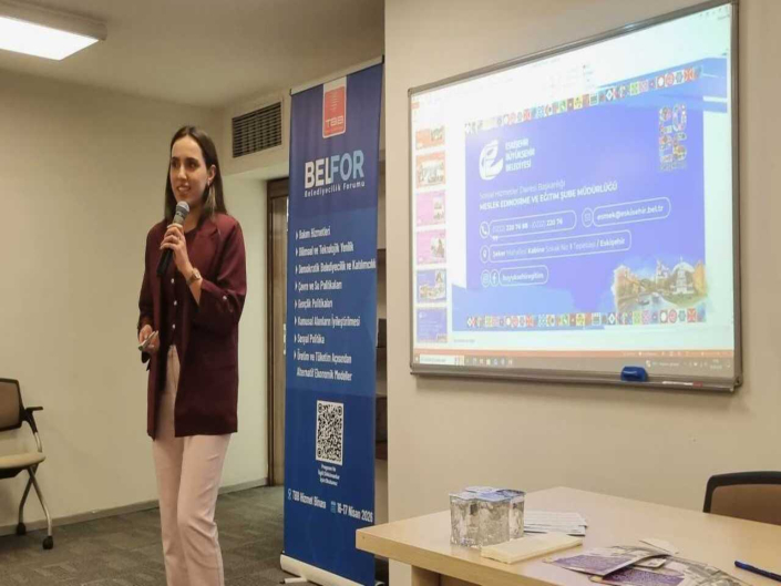 BELFOR Belediyecilik Forumu'nda sunum yapan kadın konuşmacı ve arkasındaki projeksiyon ekranı.