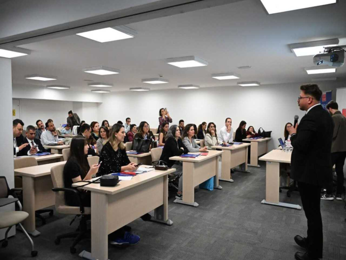Bir eğitim salonunda konuşmacıyı dinleyen katılımcılar ve sunum yapan takım elbiseli erkek konuşmacı.