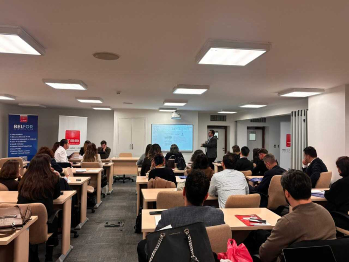 Aydınlık bir seminer salonunda, sunum yapan bir konuşmacıyı ve masalarında oturarak sunumu dinleyen katılımcıları gösteren fotoğraf.