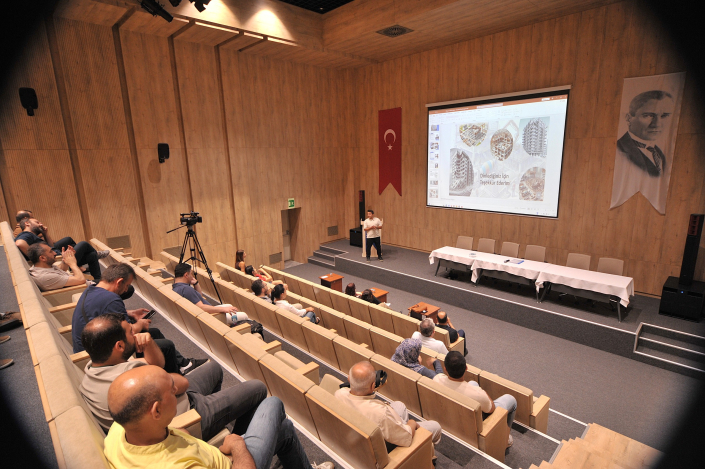 Bir seminer salonunda, duvarda Türk bayrağı, Atatürk posteri ve sahnede sunum yapan bir kişinin yer aldığı geniş, ahşap panelli bir konferans salonu ve sunumu dinleyen katılımcıların görüntüsü.