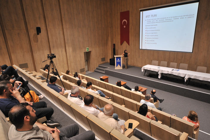 Ahşap panelli bir seminer salonunda, sahnedeki büyük ekranda 'AFET PLANI' başlıklı sunum maddelerini anlatan bir konuşmacı ve onu dinleyen katılımcılar.
