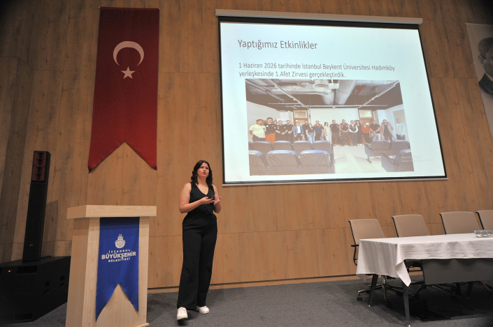 İstanbul Büyükşehir Belediyesi kürsüsünün yanında, projeksiyondaki 'Yaptığımız Etkinlikler' başlıklı sunum eşliğinde konuşma yapan bir kadın.