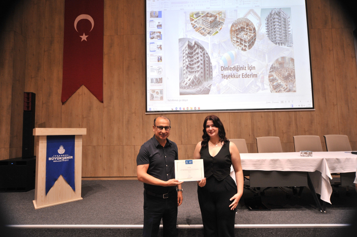 Bir seminer salonunda, arkalarında 'Dinlediğiniz İçin Teşekkür Ederim' yazılı projeksiyon perdesi önünde yan yana durarak sertifika tutan bir erkek ve bir kadın.