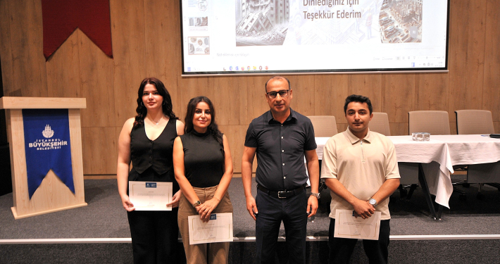Bir seminersalonunda, arkalarında 'Dinlediğiniz İçin Teşekkür Ederim' yazılı projeksiyon perdesi önünde yan yana durarak sertifikalarıyla poz veren üç katılımcı ve bir yetkili.