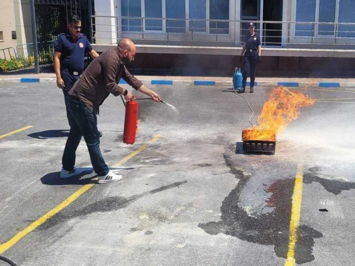 Gündüz vakti bir otoparkta yangın söndürme eğitimi. Bir personel, yangın tüpüyle küçük bir ateşe müdahale ediyor; üniformalı bir itfaiyeci onu izliyor, arka planda bir başka itfaiyeci ve bir bina var.