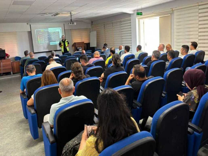 Bir eğitim salonunda, projeksiyon perdesi önünde sunum yapan yelekli eğitmeni dinleyen mavi koltuklarda oturan katılımcılar.