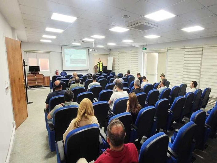 Eğitim salonunda, projeksiyon perdesi önünde sunum yapan yelekli eğitmeni dinleyen dinleyicilerin geniş açıdan görünümü.