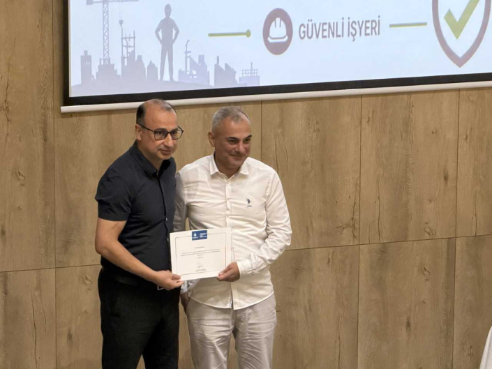 Görsel, bir İş Sağlığı ve Güvenliği Seminerine ait eğitimi sonunda belge takdimi anını göstermektedir. Arka planda ahşap panellerle kaplı bir duvar ve üzerinde bir projeksiyon perdesi bulunuyor. Perdenin görünen alt kısmında "Güvenli İşyeri" yazısı mevcut.