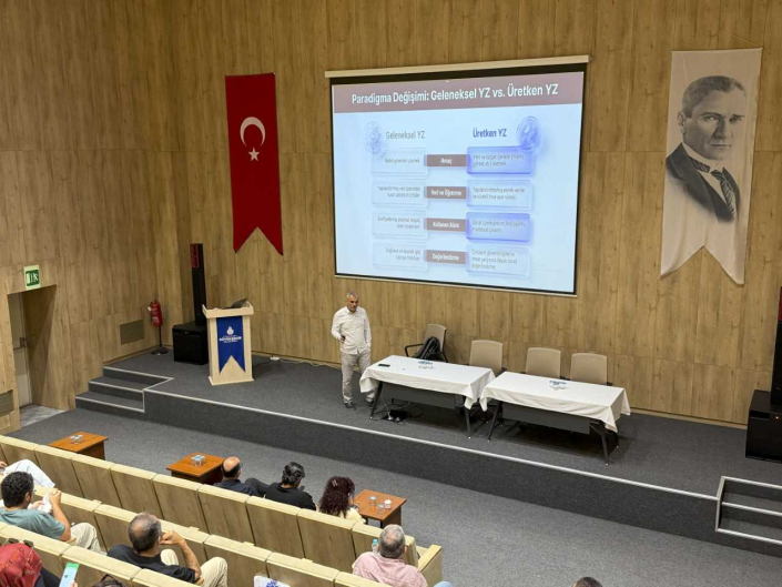 Görsel, bir konferans salonunda "Paradigma Değişimi: Geleneksel YZ vs. Üretken YZ" başlıklı sunumu aktaran bir konuşmacıyı ve dinleyicileri göstermektedir.