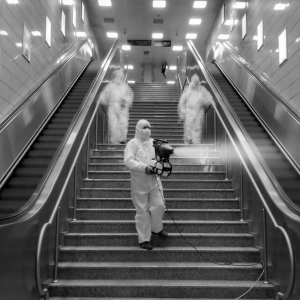 Bu siyah-beyaz fotoğraf, iki yürüyen merdiven arasındaki bir metro istasyonu merdivenini gösteriyor. Tam koruyucu giysili ve maskeli üç kişi merdivenleri temizliyor. Ön plandaki kişi elinde tuttuğu bir cihazla merdivenlere sis püskürtüyor. Diğer iki kişi arkada ve bulanık görünüyorlar.
