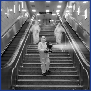 Bu siyah-beyaz fotoğraf, iki yürüyen merdiven arasındaki bir metro istasyonu merdivenini gösteriyor. Tam koruyucu giysili ve maskeli üç kişi merdivenleri temizliyor. Ön plandaki kişi elinde tuttuğu bir cihazla merdivenlere sis püskürtüyor. Diğer iki kişi arkada ve bulanık görünüyorlar.