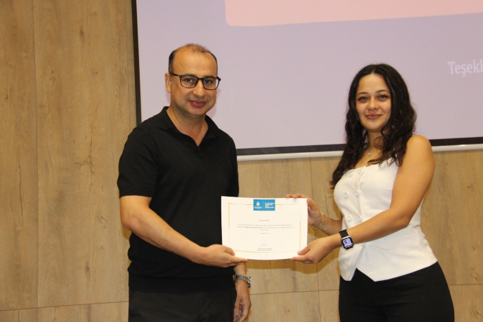 Görselde, bir projektör ekranının ön kısmında duran iki kişi görülüyor. Solda duran ve siyah polo yaka tişöert giymiş bir erkek, sağda ise beyaz renk üst ve siyah renk kıyafet gyimiş alt bir kadın yer alıyor. İkisi de gülümseyerek kameraya bakıyor ve birlikte bir sertifika tutuyorlar.