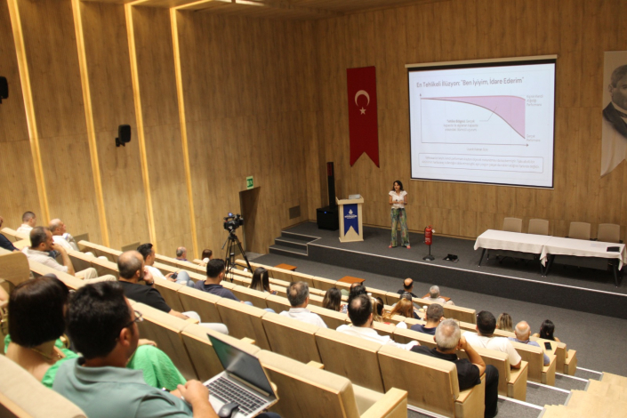 Modern tasarımlı, ahşap panel kaplamalı geniş bir seminer salonunda, dinleyicilere sunum yapan bir kadın konuşmacı ve duvara yansıtılmış sunum slaytı. Slaytın üst başlığında "En Tehlikeli İllüzyon: 'Ben İyiyim, İdare Ederim'" yazmaktadır.