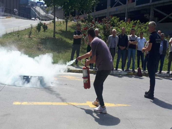 Yangın tatbikatında, bir adamın söndürme tüpüyle dumanlar yükselen alana müdahalesini izleyen kalabalık grup ve itfaiye görevlisi.