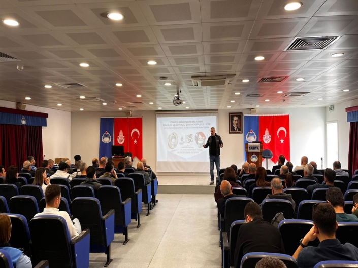 Konferans salonunda, projeksiyon perdesinin önünde ayakta sunum yapan bir eğitmen ve salonu dolduran dinleyiciler. Sunum perdesinin solunda Atatürk portresi, Türk Bayrağı, İBB Flaması ve Zabıta Flaması, sağında ise Türk Bayrağı, İBB Flaması ve Zabıta Flaması yer alıyor.